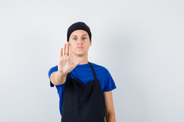 Młody kucharz mężczyzna w t-shirt, fartuch pokazując gest stop i patrząc poważnie, widok z przodu.