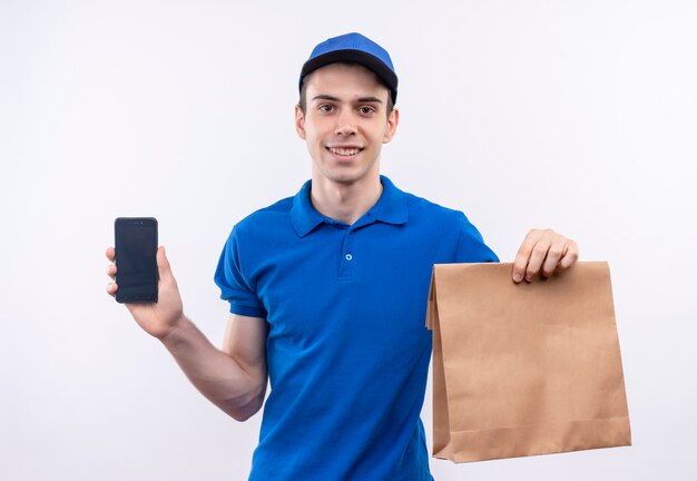 Młody kurier w niebieskim mundurze i niebieskiej czapce szczęśliwie trzyma torbę i telefon