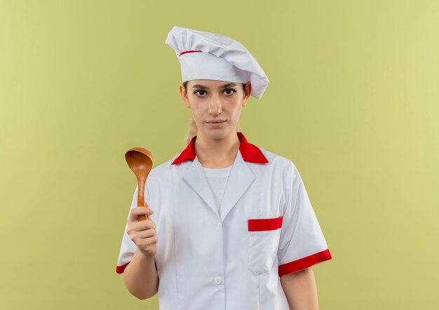 Młody ładny kucharz w mundurze szefa kuchni trzymając łyżkę i patrząc na białym tle na zielonym tle