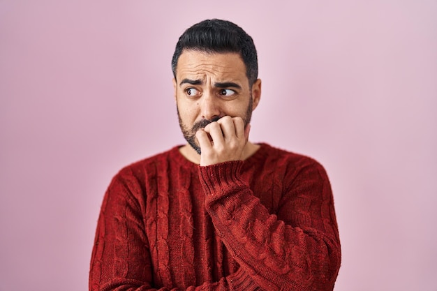 Młody latynos z brodą, ubrany w luźny sweter na różowym tle, wyglądający na zestresowanego i zdenerwowanego z rękami na ustach obgryzającymi paznokcie problem lęku