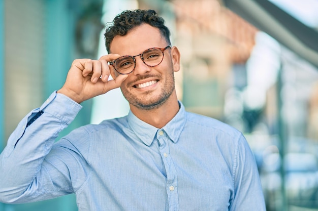 Bezpłatne zdjęcie młody latynoski biznesmen uśmiecha się szczęśliwy, dotykając okularów w mieście.