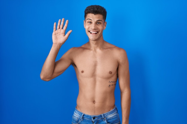 Bezpłatne zdjęcie młody latynoski mężczyzna stojący bez koszuli na niebieskim tle, rezygnujący z witania się szczęśliwym i uśmiechniętym przyjaznym gestem powitalnym