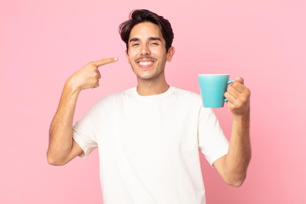 Młody latynoski mężczyzna uśmiechający się pewnie, wskazując na swój szeroki uśmiech i trzymający kubek z kawą
