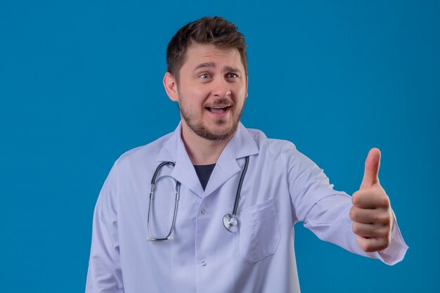 Młody lekarz mężczyzna ubrany w biały fartuch i stetoskop pokazujący kciuki do góry gest z radosną buźką na odosobnionym niebieskim tle 1