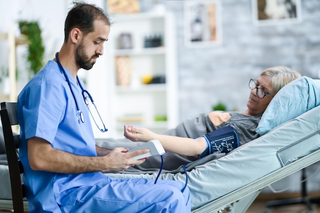 Młody lekarz ze stetoskopem w domu opieki robi badanie ciśnienia krwi starej kobiety za pomocą urządzenia cyfrowego.