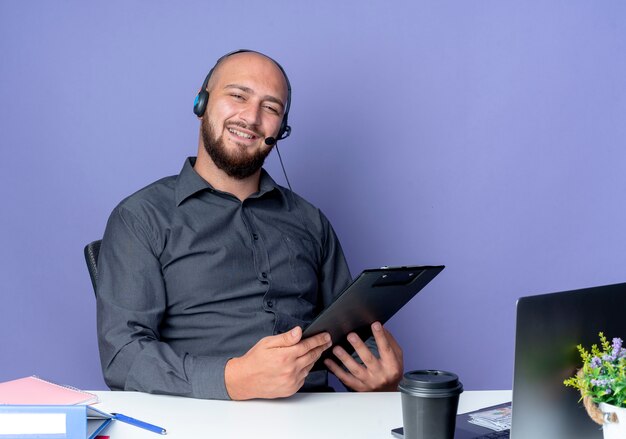 Młody łysy mężczyzna call center sobie zestaw słuchawkowy siedzi przy biurku z narzędzi pracy na białym tle na fioletowym tle