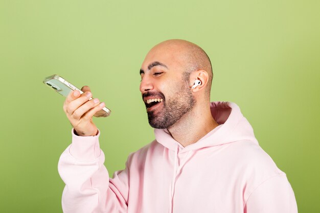 Młody łysy mężczyzna kaukaski w różowej bluzie z kapturem na białym tle, trzymaj telefon szczęśliwy pozytywny nagrywanie wiadomości audio