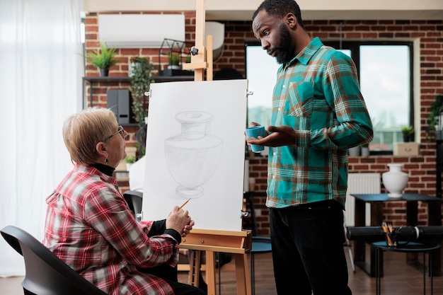 Młody malarz nauczyciel wyjaśniający szkic ilustracji starszej kobiecie pracującej razem przy rysowaniu graficznym w studio kreatywności. Zróżnicowany zespół rysujący model wazonu na płótnie uczący się nowych umiejętności artystycznych