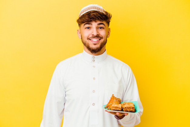 Młody marokański mężczyzna ubrany w typowy strój arabski jedzący arabskie słodycze na żółtej ścianie szczęśliwy, uśmiechnięty i wesoły.