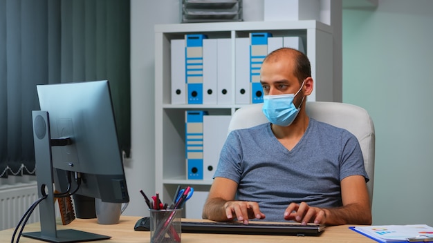 Młody menedżer z maską ochronną pracuje samotnie w biurze podczas dystansu społecznego. Przedsiębiorca w nowym, normalnym, osobistym miejscu pracy, pisanie na klawiaturze komputera, patrząc na pulpit