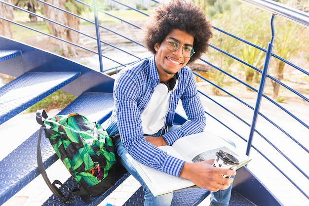 Młody męski uczeń siedzi na schody z plecakiem; książka i filiżanka kawy na wynos