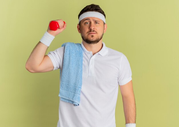 Młody mężczyzna fitness w białej koszuli z pałąkiem na głowę i ręcznikiem na ramieniu, trzymając hantle, robi ćwiczenia patrząc pewnie stojąc na oliwkowym tle