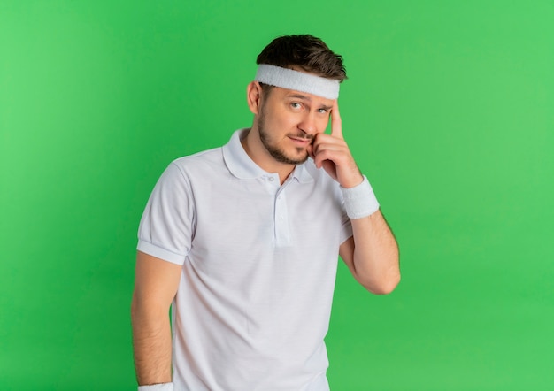 Młody mężczyzna fitness w białej koszuli z pałąkiem na głowę, patrząc do przodu z zamyślonym wyrazem myśli stojącej nad zieloną ścianą