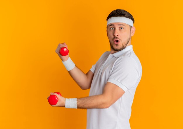 Młody mężczyzna fitness w białej koszuli z pałąkiem na głowę pracy z hantlami patrząc do przodu uśmiechnięty stojący nad pomarańczową ścianą