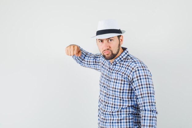 Młody mężczyzna grozi pięścią w koszuli w kratę, kapeluszu i wygląda wściekle. przedni widok.
