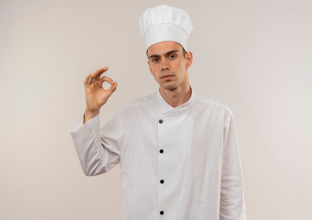 młody mężczyzna kucharz ubrany w mundur szefa kuchni pokazujący gest okey na odosobnionej białej ścianie z miejsca na kopię