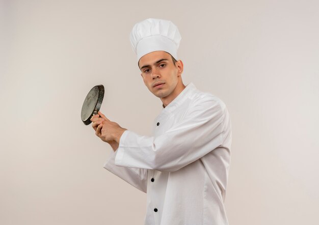 młody mężczyzna kucharz ubrany w mundur szefa kuchni trzymając patelnię wokół ramienia na odizolowanej białej ścianie