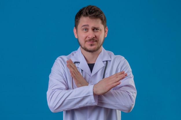 Młody mężczyzna lekarz ubrany w biały fartuch i stetoskop stojący ze skrzyżowanymi rękami gest zatrzymania na na białym tle niebieskim tle