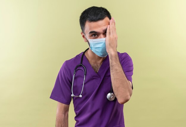 Młody Mężczyzna Lekarz Ubrany W Fioletową Odzież Chirurga I Stetoskop Maska Medyczna Zakryte Oko Ręką Na Izolowanej Zielonej ścianie