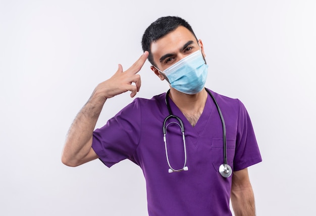 młody mężczyzna lekarz ubrany w fioletową odzież chirurga i stetoskop medyczną maskę pokazującą gest pistoletu na odizolowanej białej ścianie