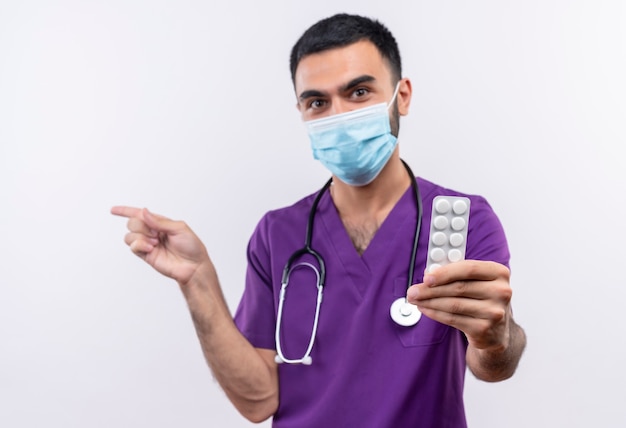 młody mężczyzna lekarz ubrany w fioletową odzież chirurga i stetoskop medyczną maskę, wyciągając tabletki do kamery wskazuje na bok na odosobnionej białej ścianie