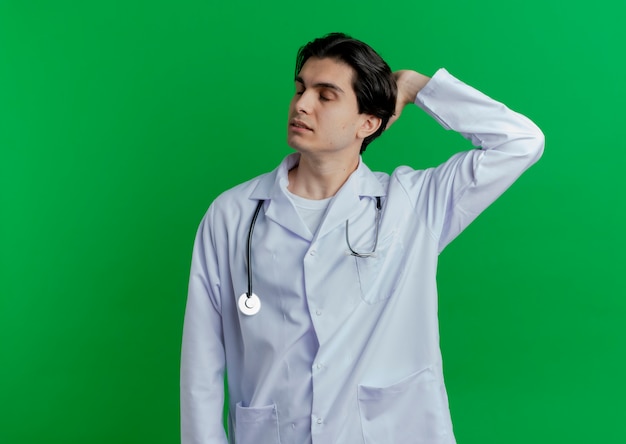Młody mężczyzna lekarz ubrany w medyczną szatę i stetoskop trzymając rękę za głową z zamkniętymi oczami na białym tle