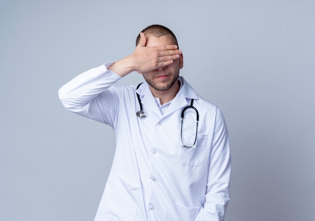 Młody mężczyzna lekarz ubrany w medyczną szatę i stetoskop wokół szyi, zamykając oczy ręką na białym tle z miejsca kopiowania