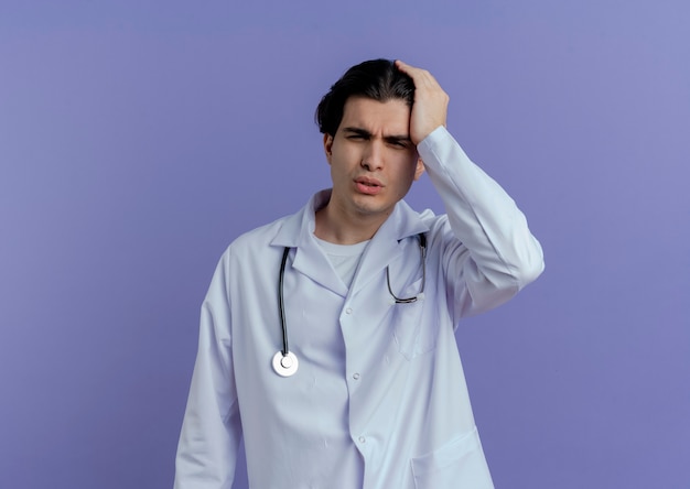 Młody mężczyzna lekarz ubrany w szlafrok medyczny i stetoskop patrząc z boku, trzymając rękę na głowie cierpiącej na ból głowy na fioletowej ścianie z miejscem na kopię
