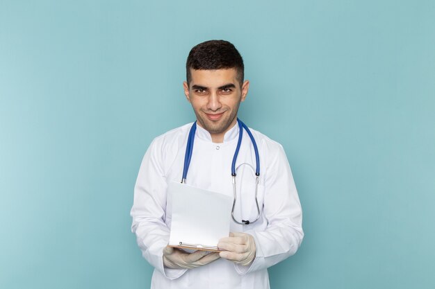 Młody mężczyzna lekarz w białym garniturze z niebieskim stetoskopem trzymając notatnik i uśmiechając się