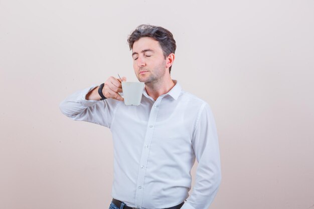 Młody mężczyzna pachnący aromatyczną herbatą w białej koszuli, dżinsach i wyglądający na zachwyconego