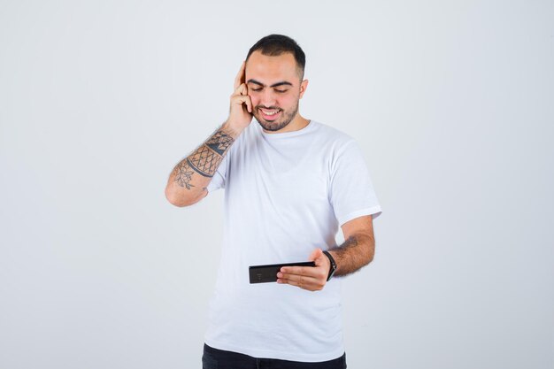 Młody mężczyzna patrzący na coś na telefonie i naciskający dłoń na ucho w białej koszulce i czarnych spodniach i wyglądający na szczęśliwego