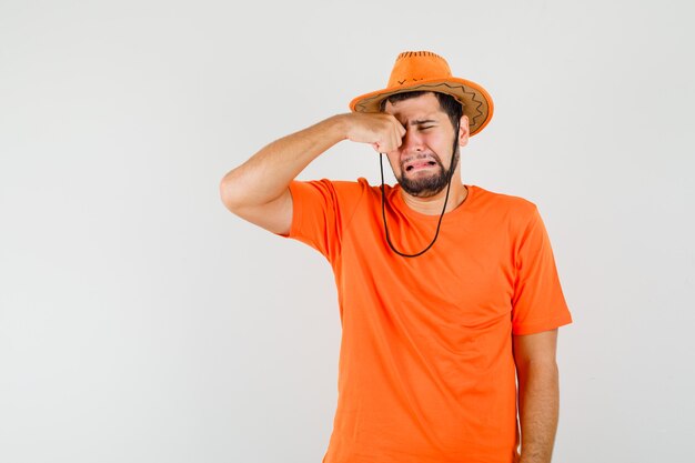 Młody mężczyzna pociera oko płacząc jak dziecko w pomarańczowej koszulce, kapeluszu i wygląda na obrażonego, widok z przodu.