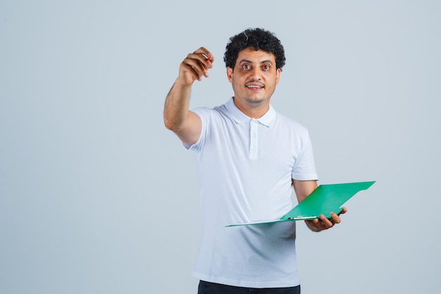Młody mężczyzna podnoszący pióro w geście eureka i trzymający notatnik w białej koszulce i dżinsach i wyglądający rozsądnie. przedni widok.