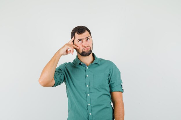 Młody mężczyzna pokazując znak v na twarzy w zielonej koszuli i patrząc zamyślony, widok z przodu.