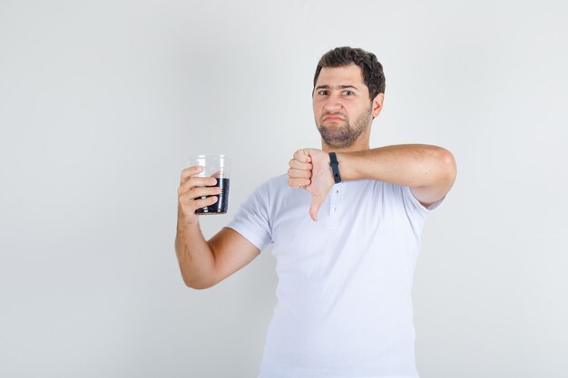 Młody mężczyzna pokazuje kciuk w dół z napojem cola w białej koszulce i wygląda smutno