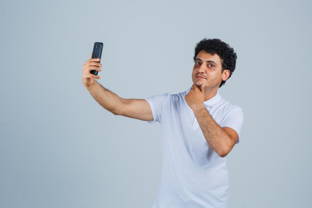 Młody mężczyzna pozuje podczas robienia selfie w białej koszulce i wygląda ładnie. przedni widok.