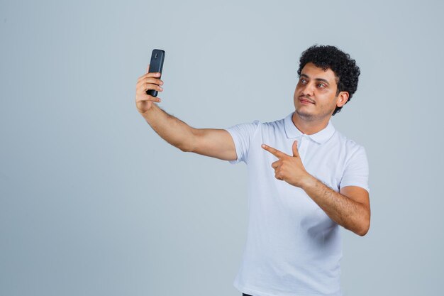 Młody mężczyzna pozuje podczas robienia selfie w białej koszulce i wygląda ładnie. przedni widok.