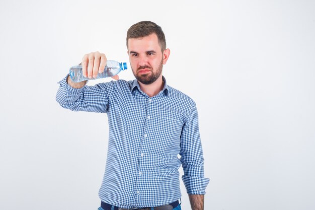 Młody mężczyzna przechylając plastikową butelkę wody w koszuli, dżinsy i niezdecydowany, widok z przodu.