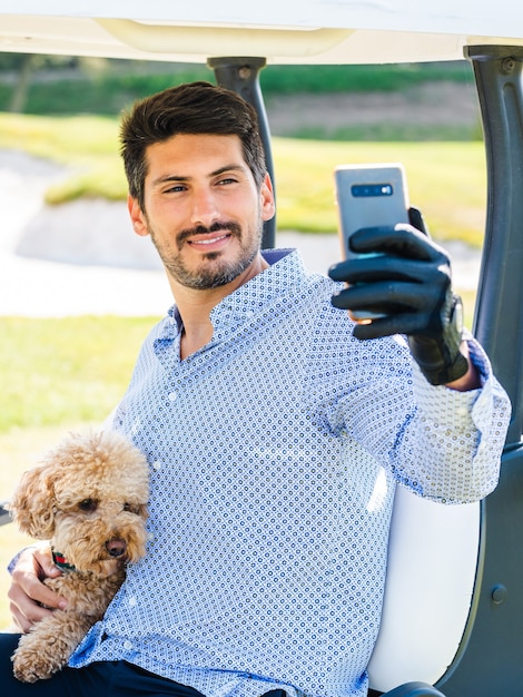 Młody Mężczyzna Rasy Kaukaskiej W Wózku Golfowym Robi Selfie Ze Swoim Szczeniakiem Goldendoodle Na Polu Golfowym