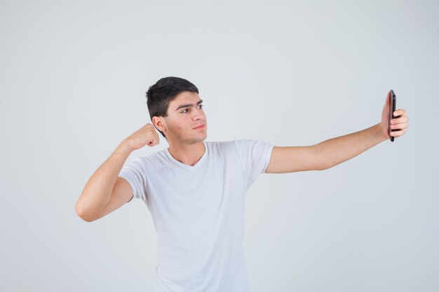 Młody mężczyzna robi selfie, pokazując mięśnie ramion w koszulce i patrząc wesoło. przedni widok.