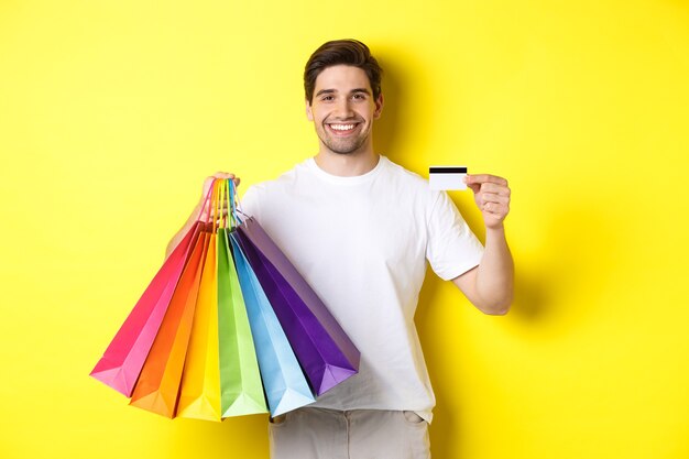 Młody mężczyzna robi zakupy na wakacje, trzymając papierowe torby i polecając bankową kartę kredytową, stojąc na żółtym tle.
