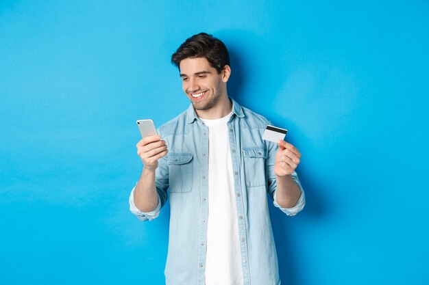 Młody mężczyzna robi zakupy online za pomocą aplikacji mobilnej, trzymając smartfon i kartę kredytową, stojąc na niebieskim tle