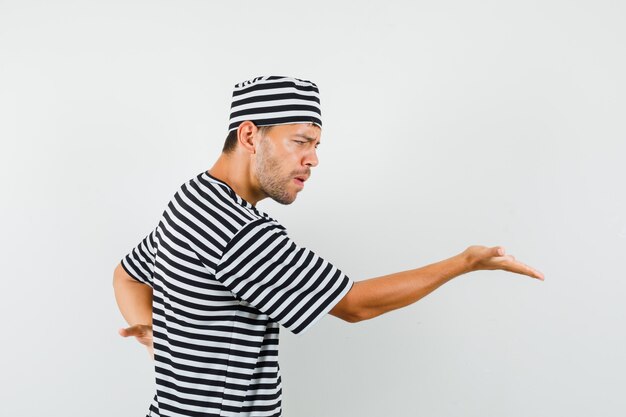 Młody mężczyzna rozciągający rękę w pytający sposób w kapelusz w paski t-shirt i patrząc zły