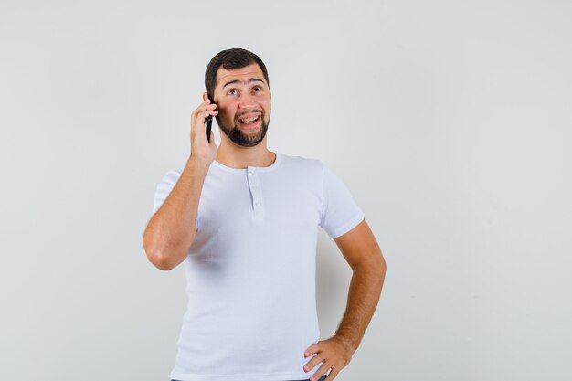 Młody mężczyzna rozmawia przez telefon w białej koszulce i patrząc rozmowny, widok z przodu.