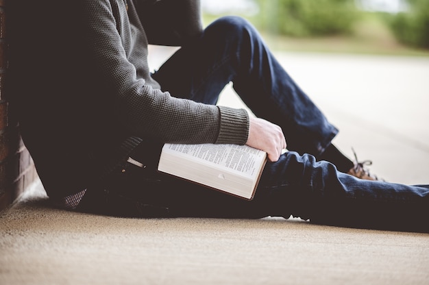 Bezpłatne zdjęcie młody mężczyzna siedzi na ziemi i trzyma biblię w dłoniach