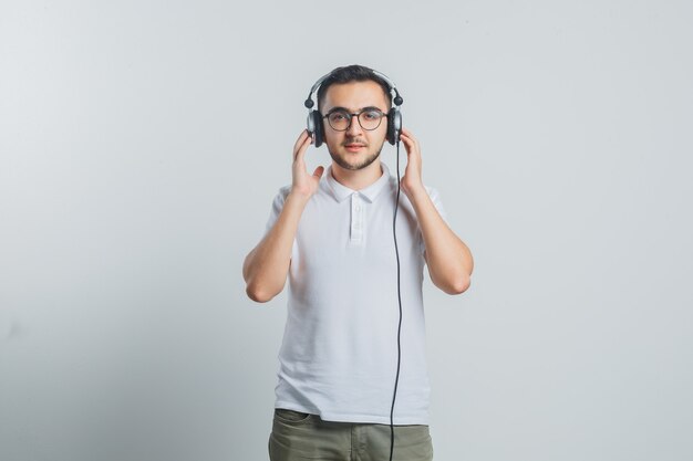 Młody mężczyzna słucha muzyki w słuchawkach w białej koszulce, spodniach i wygląda pewnie