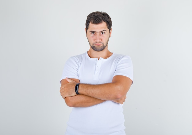 Młody mężczyzna stojący ze skrzyżowanymi rękami w białej koszulce i patrząc zamyślony