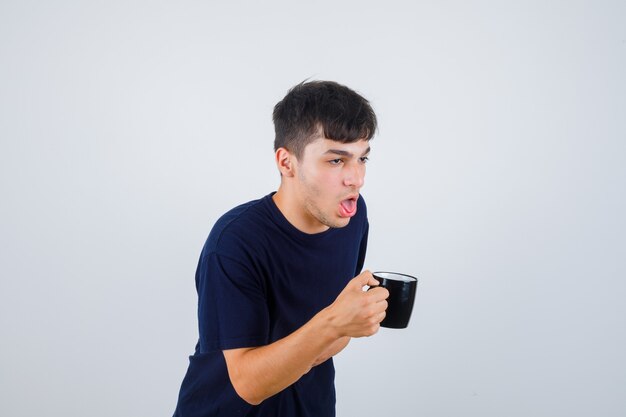Młody mężczyzna trzyma filiżankę herbaty, czując mdłości w czarnej koszulce i źle wyglądając. przedni widok.