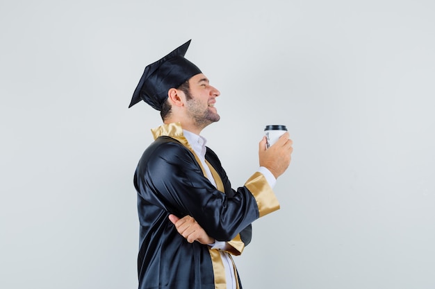 Młody mężczyzna trzyma filiżankę kawy w mundurze absolwenta i wygląda wesoło. .