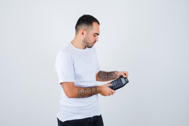 Młody mężczyzna trzyma kalkulator i robi na nim operacje w białej koszulce i czarnych spodniach i wygląda poważnie